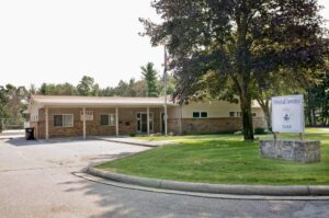 A front photo of Whitehall Township Hall.  The building is brown and tan with a green lawn and vehicle parking in front of it.